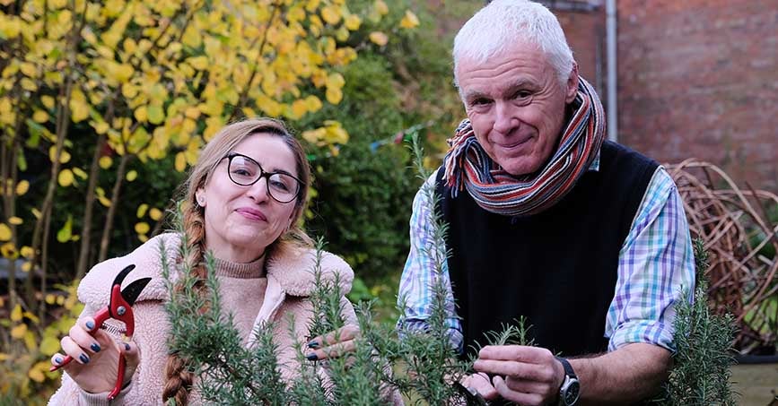 Man and woman stood in a garden, holding on to a plant, smiling. 
