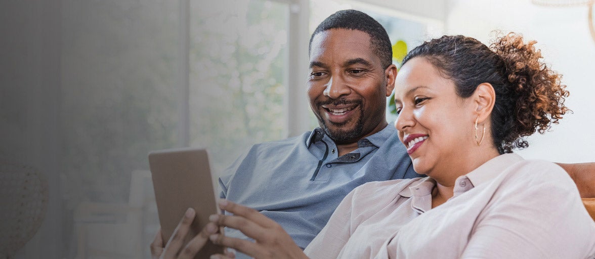Man and woman sat together on the sofa, looking at a mobile phone smiling. 