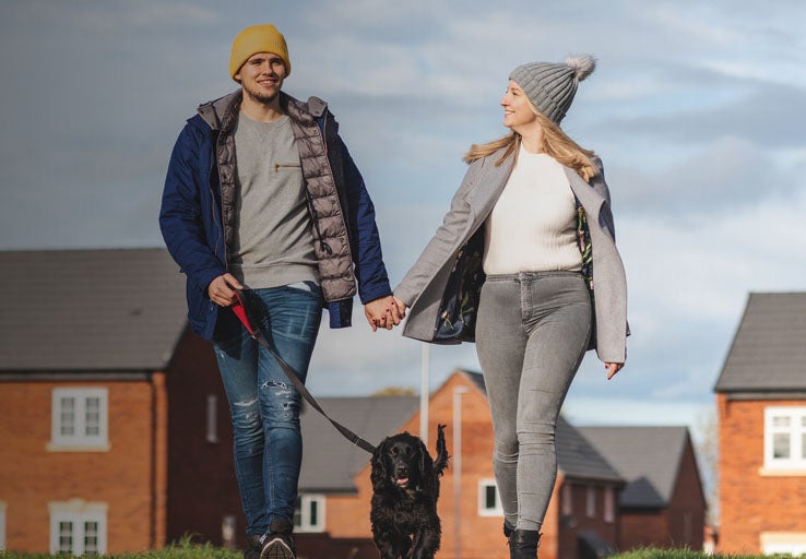 A couple walking their dog with houses in the background. 