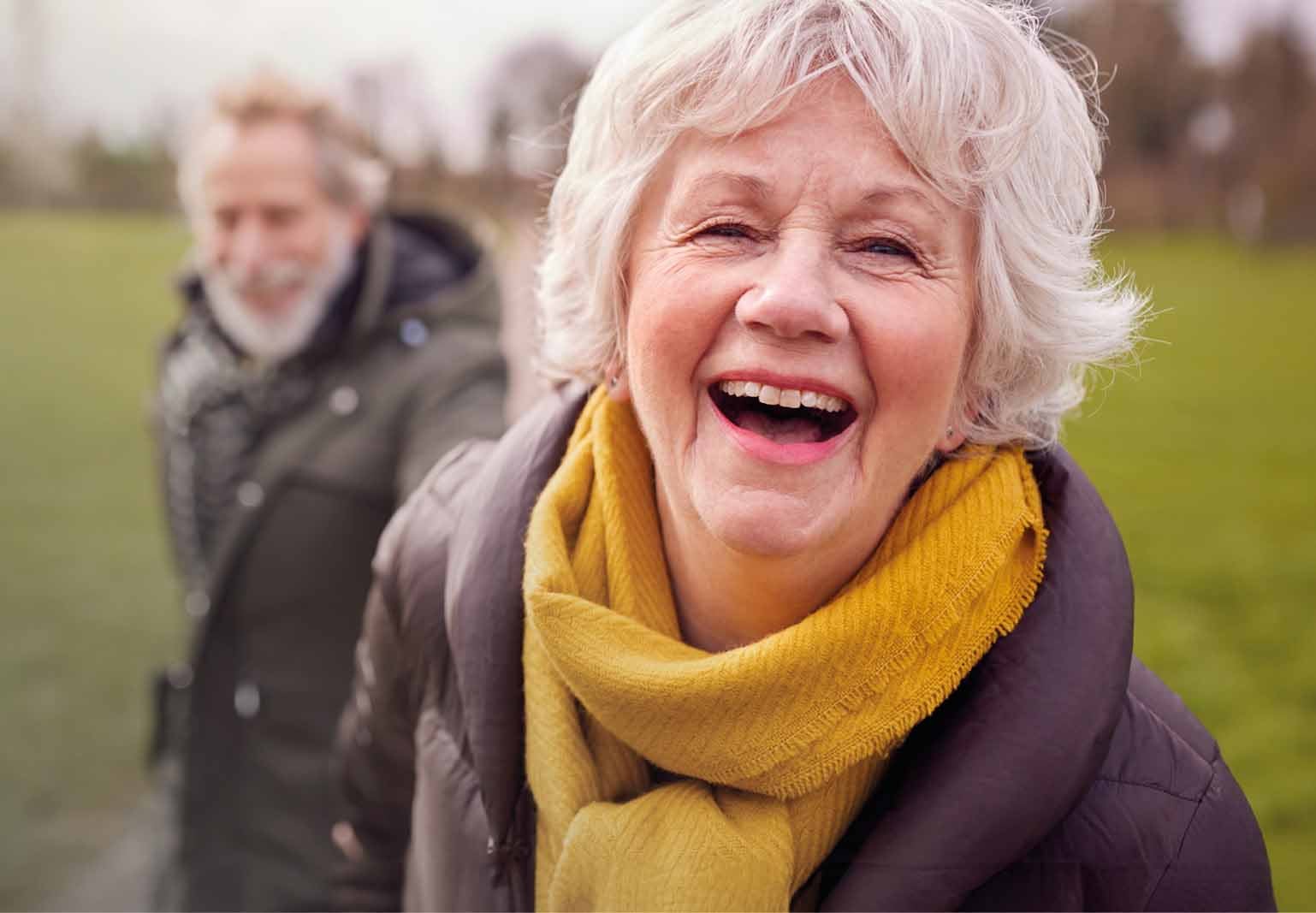 An older lady laughing into the camera with her husband in the background.