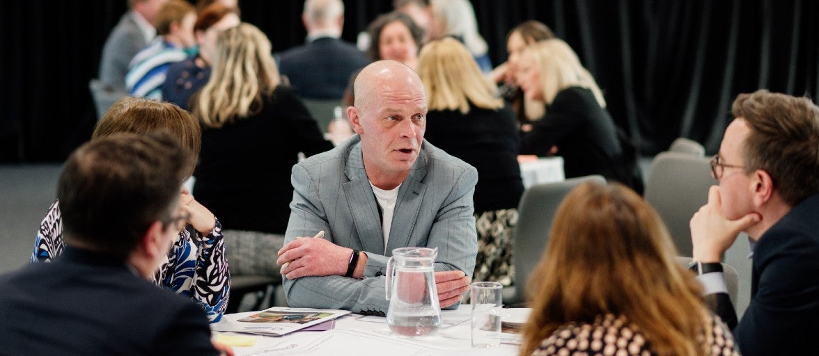 A group of people sat around a table listening to the man in the middle speak. In the background there is another group discussion at a different table.
