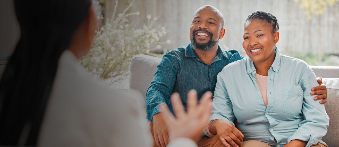 A husband and wife sat on a sofa with arms around each other smiling at their financial adviser. 