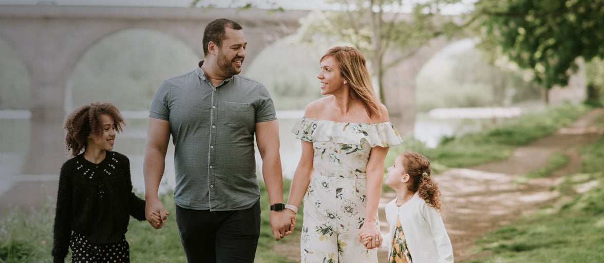 A family comprising of 2 adults and two children hold hands while walking by the river in Hexham
