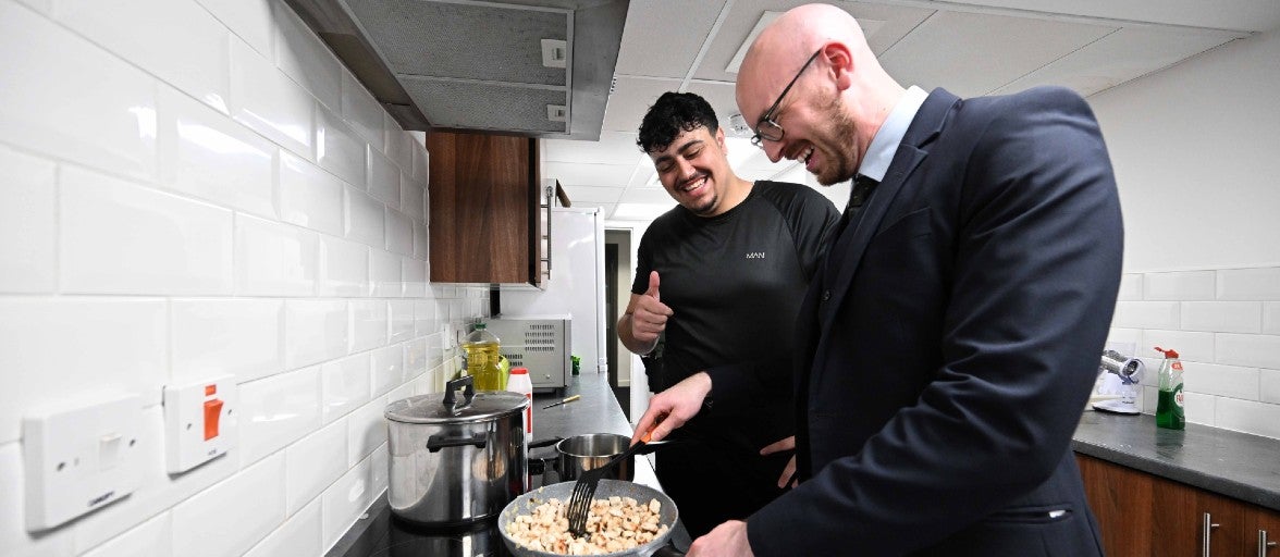 Two men stood smiling beside a kitchen stove as one of the men stirs some food in a saucepan.