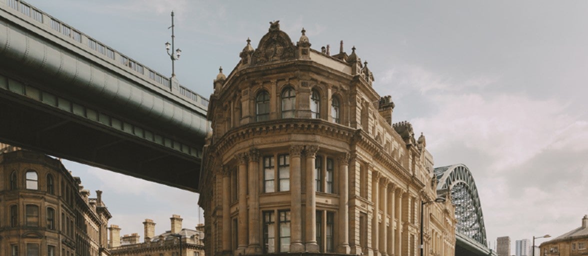 People walking down a street in front of a large building with the Tyne Bridge in the background.