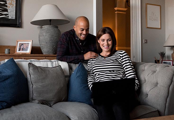 Man and woman sat in living room looking at a laptop smiling.