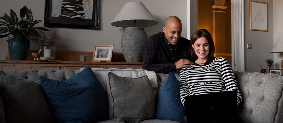 Couple at home in their living room, wife sat on the sofa with a laptop resting on her knees, whilst husband bends down to look over her shoulder. 