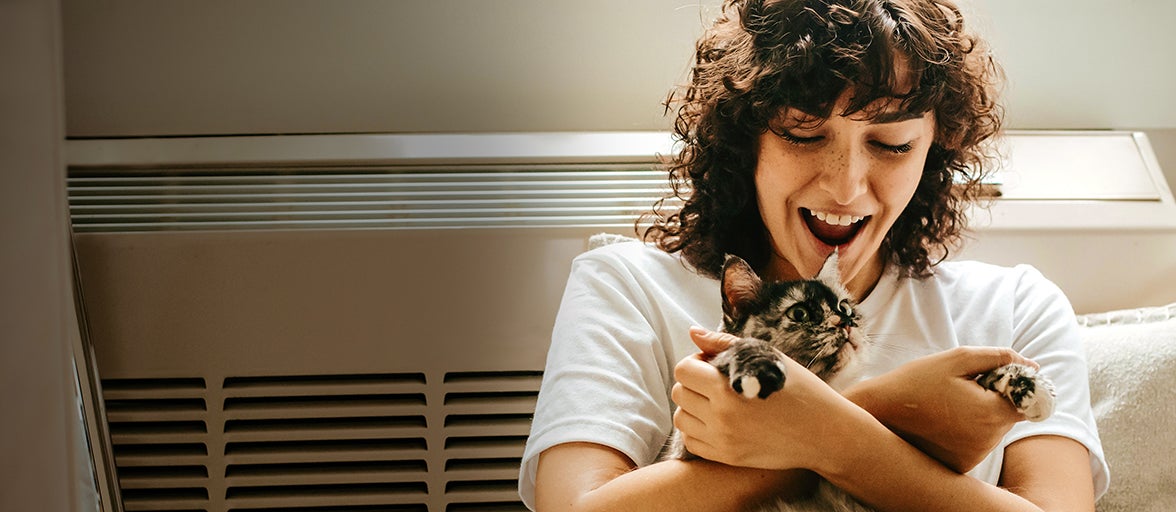 A lady smiles as she cuddles a cat as she rests against a radiator.