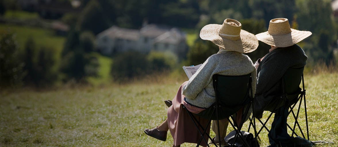 Two elderly people sat on chairs in a field overlooking countryside. Both wearing wide brim straw hats, lady is holding a crossword puzzle book. 