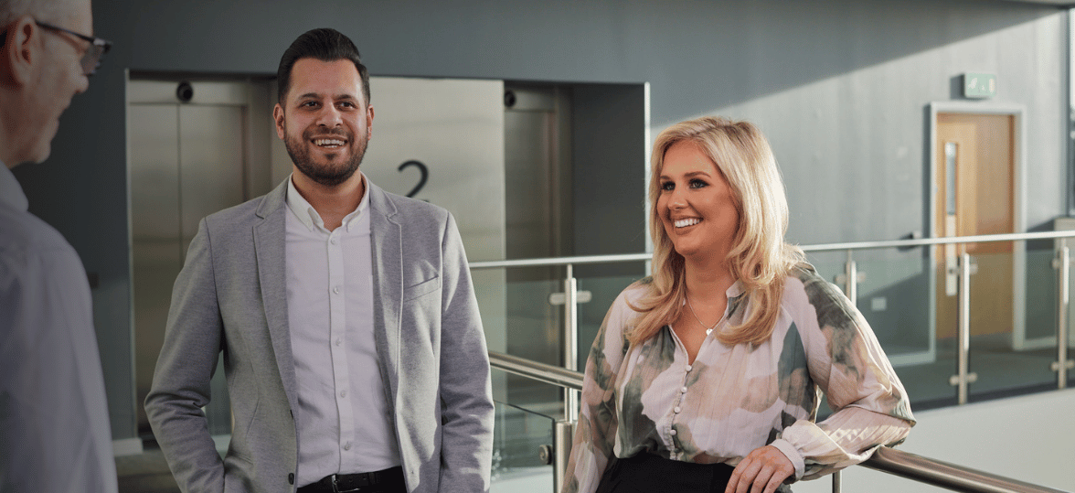Two people standing in an office corridor, smiling and looking at each other.