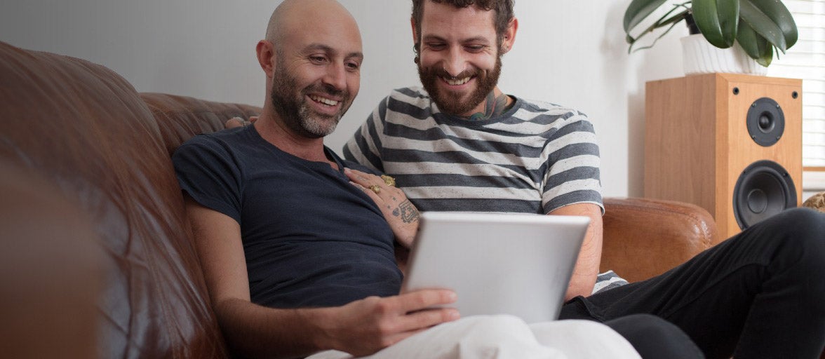 A couple sat on their sofa looking at their laptop, smiling. 