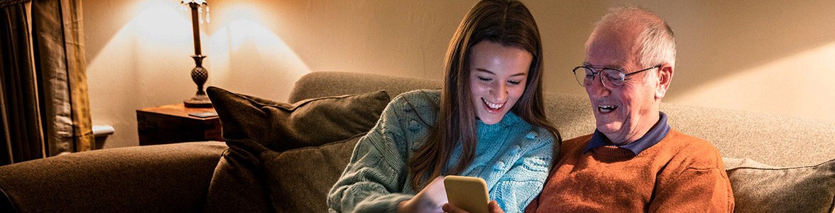 Senior man sat next to his teenage granddaughter on the sofa, looking at a mobile phone.