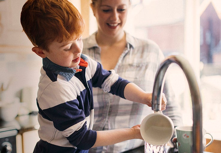 Little boy pouring water out of a cup in the kitchen, his mum stands behind him. 