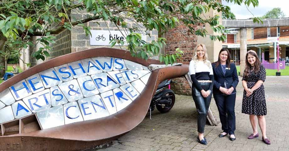Three ladies stood outside of Henshaws & Crafts Centre. In the foreground on the left-hand side, is the large sign for the Centre and the ladies are stood to the right. The Centre is visible in the background.