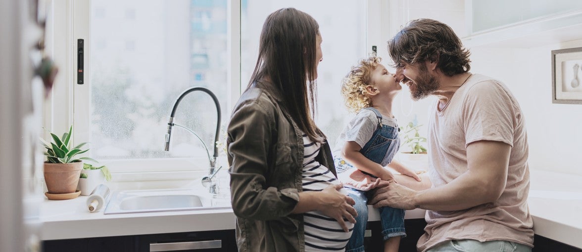 A pregnant lady, father and a young child cuddle in the kitchen.