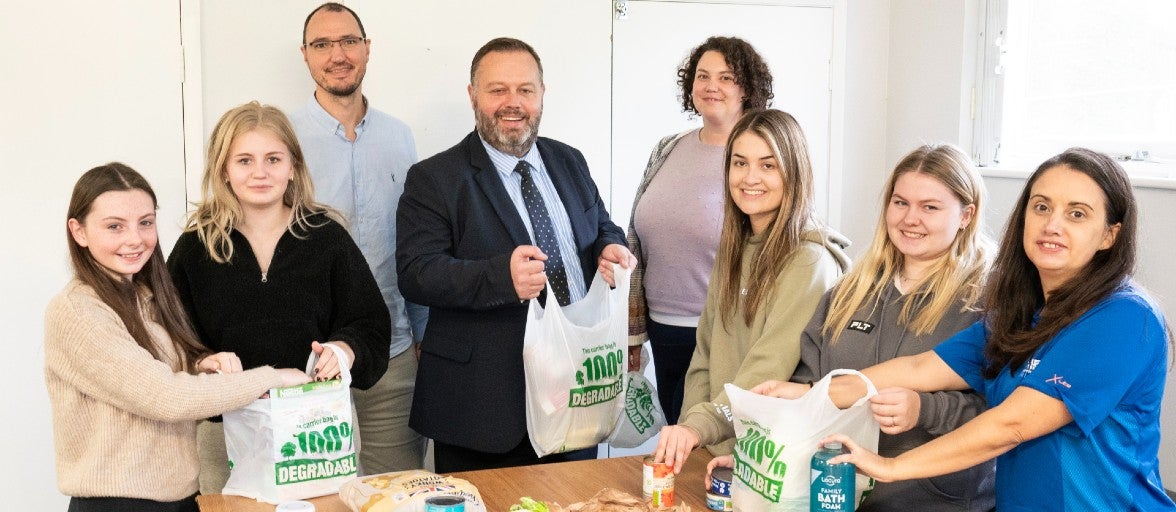 A group of people stood holding shopping bags full of food, behind a table covered in tins of food, jars of sauces, and loose fruit and vegetables.