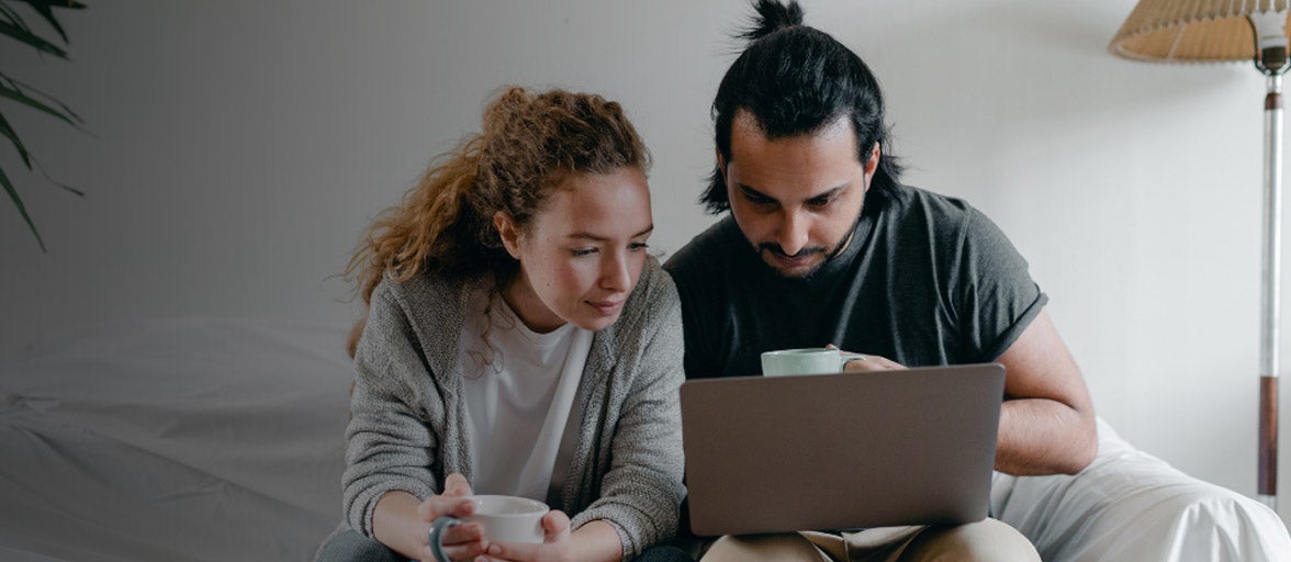 couple sat on sofa with coffee cups looking at their laptop