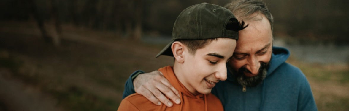 Father and son standing together smiling, Father's arm around Son's shoulder, heads together. 