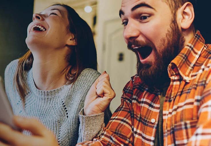Young couple, a man and woman, sat together looking at their mobile phone celebrating.