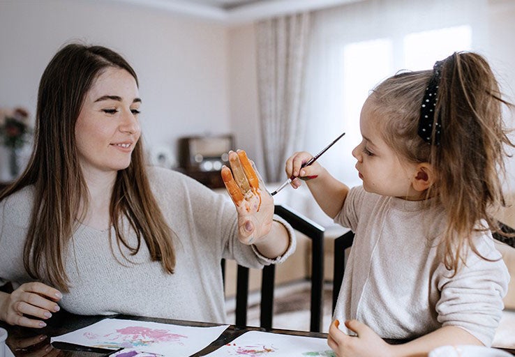 Mother and daughter sat in their home and the little girl paints orange paint onto her mothers hand. 
