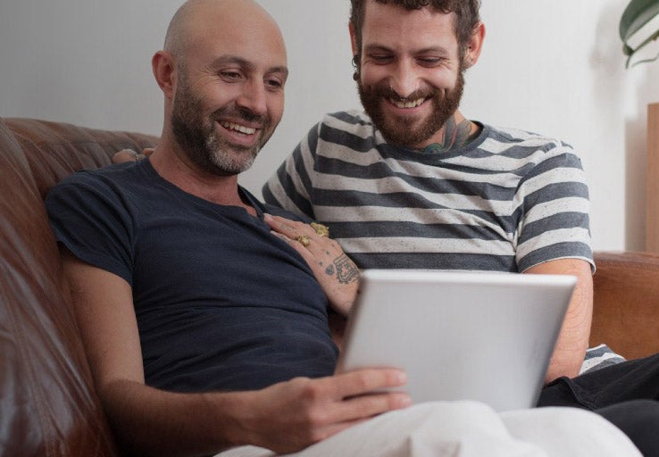 A couple sat on their sofa looking at their laptop, smiling. 