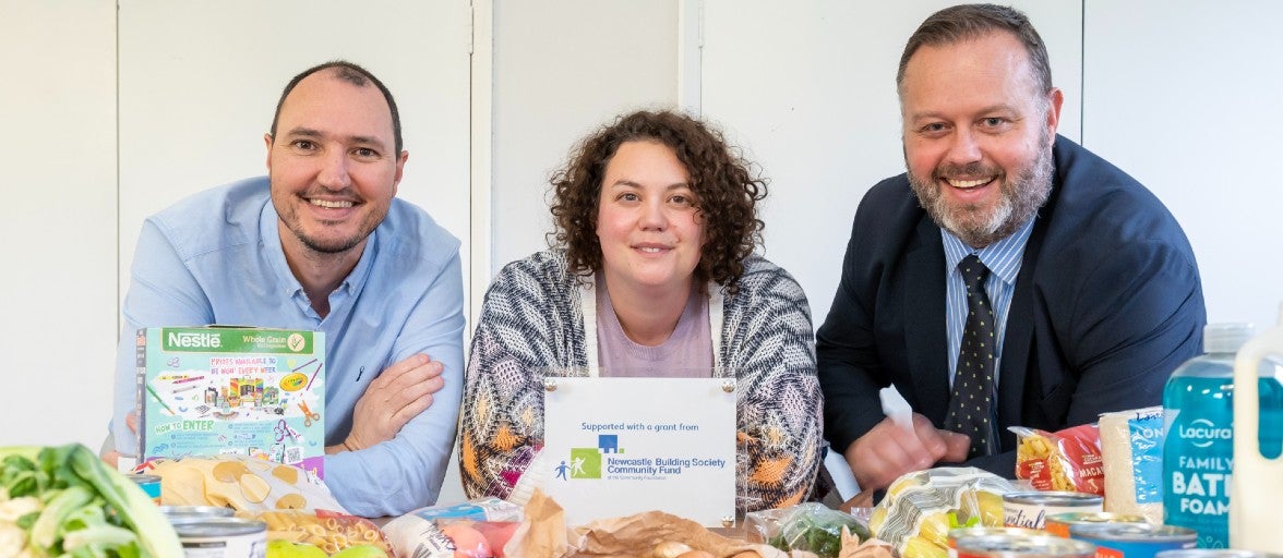 Two men and a woman smiling as they lean over a table covered in loose vegetables and assorted boxes, tins and packets of food.