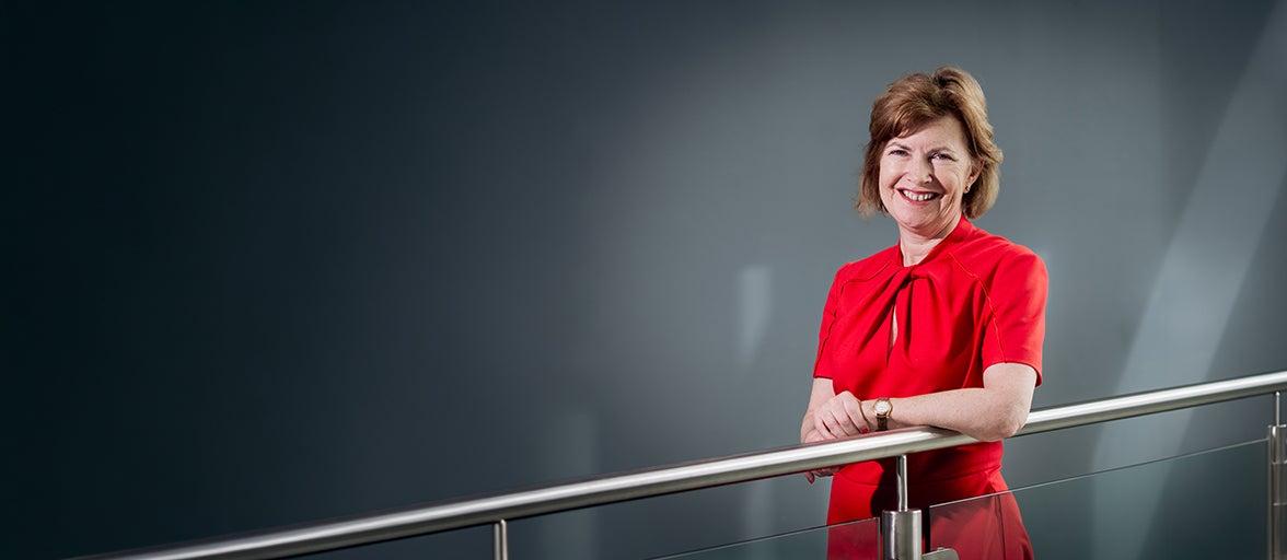 Lucy Winskell stood smiling wearing a red dress with a grey background. 