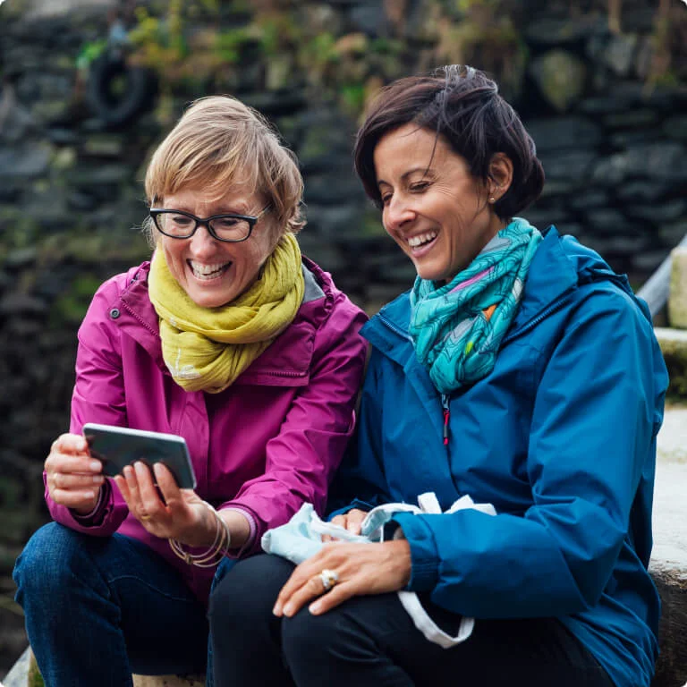 2 friends sitting on some steps while laughing at a phone. 