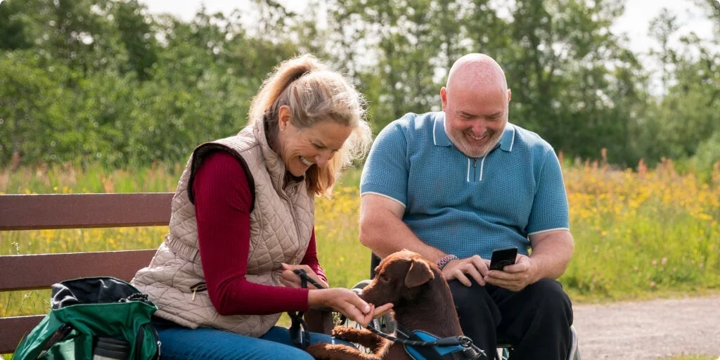 Woman sitting on bench feeding a dog. Man beside her is on the phone. 