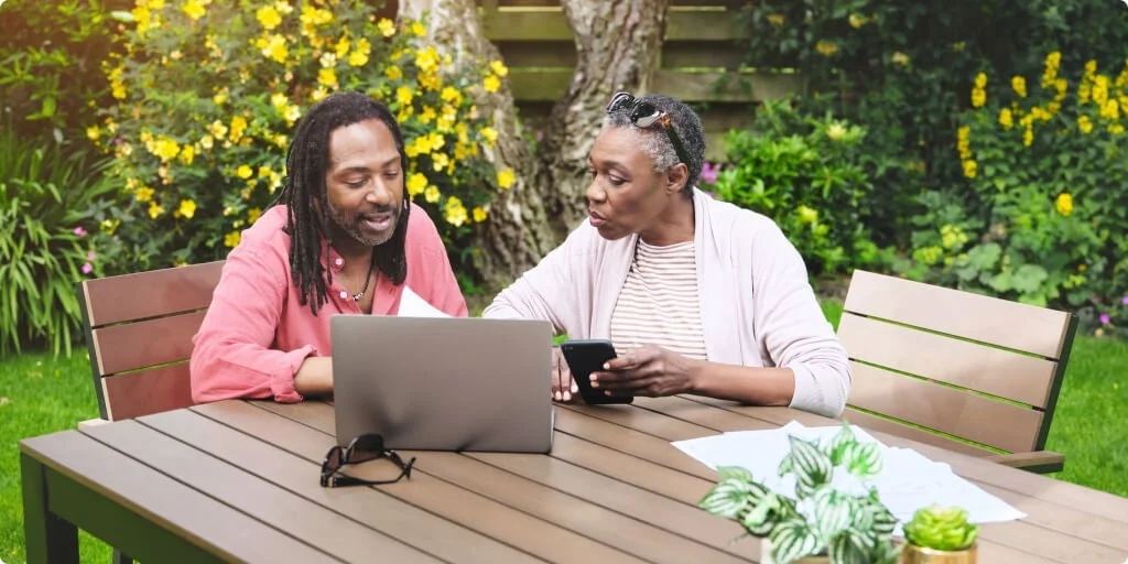 2 friends sit at a garden table looking at a phone and a laptop.