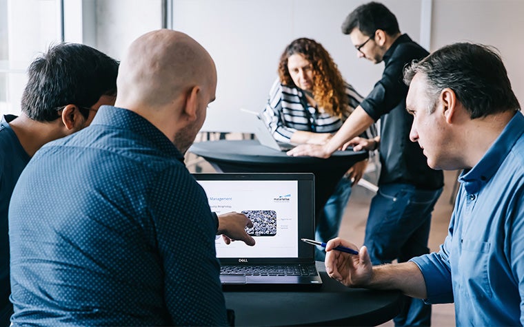 Group of three men standing at a table, looking at a laptop, with a man and a woman standing at a table in the background