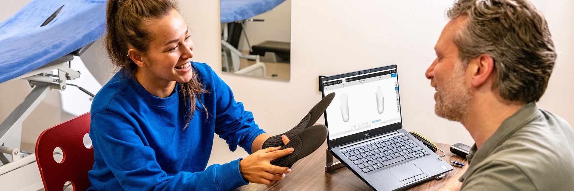 A woman holding a pair of custom-made phits orthotics sitting at a desk in a clinic opposite a male podiatrist.