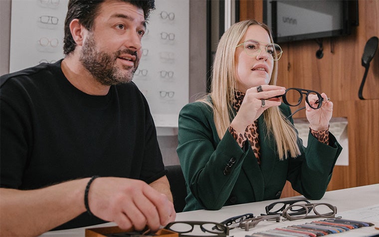 Zwei Personen halten 3D-gedruckte Brillengestelle an einem Tisch mit vielen 3D-gedruckten Gestellen hoch