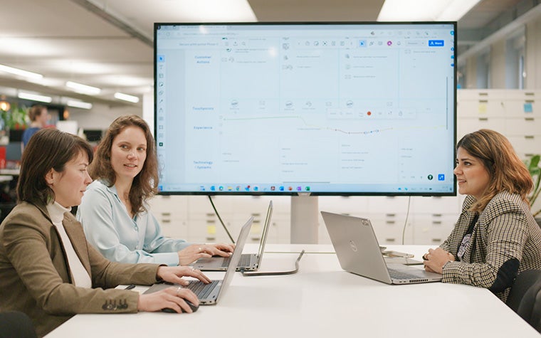 Three women sitting at a table in a meeting on their laptops with a large screen showing a planning tool