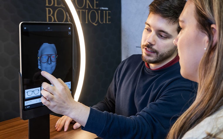 A man and a woman using the Eyewear Fitting Suite on an iPad. The screen shows a 3D model of a head wearing generated 3D-printed eyewear.