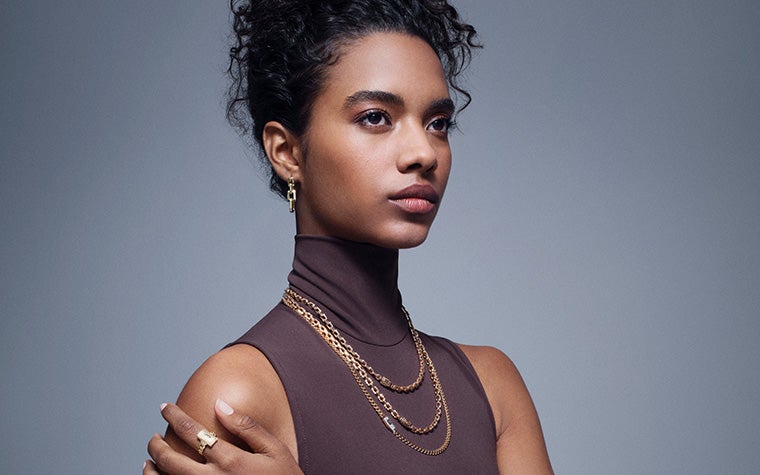 ノースリーブのシャツを着て右を見つめる女性。手は肩に触れている。首には細い金の鎖が3本、人差し指には金の指輪。