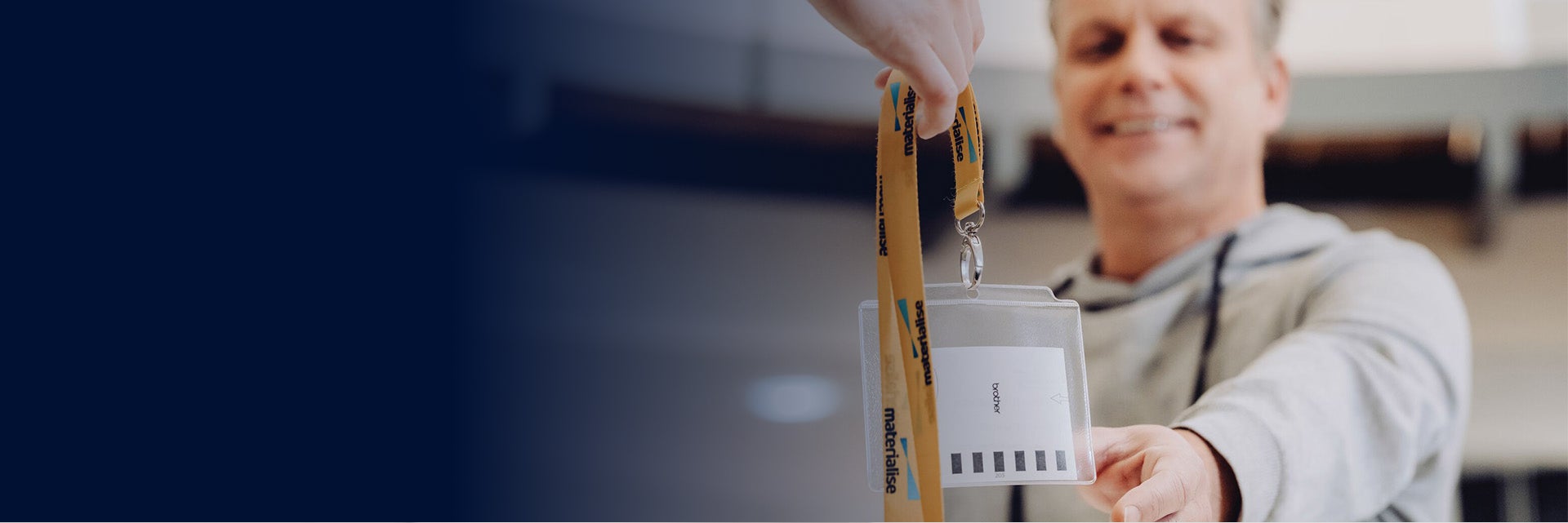 An image of a man holding out his hand reaching for a materialise visitor's badge. The badge has a yellow lanyard with the Materialise company name and logo.