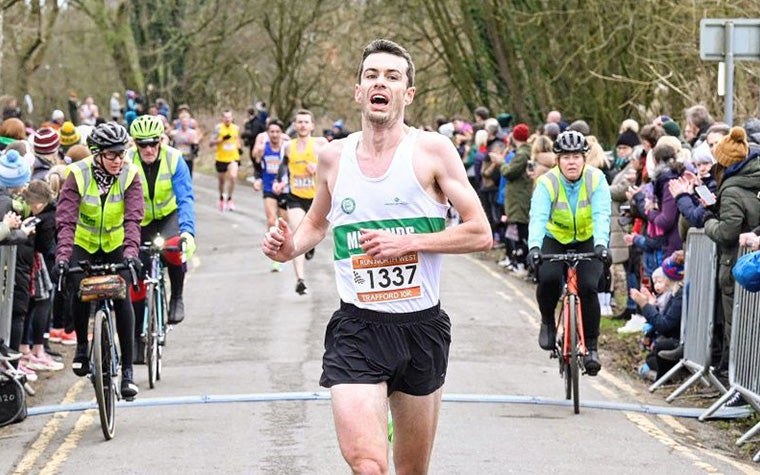 Distance runner Dan Studley crosses the finish line in the 2023 Trafford 10k, followed by three people on bicycles wearing high-vis jackets. A crowd of spectators stand on either side.