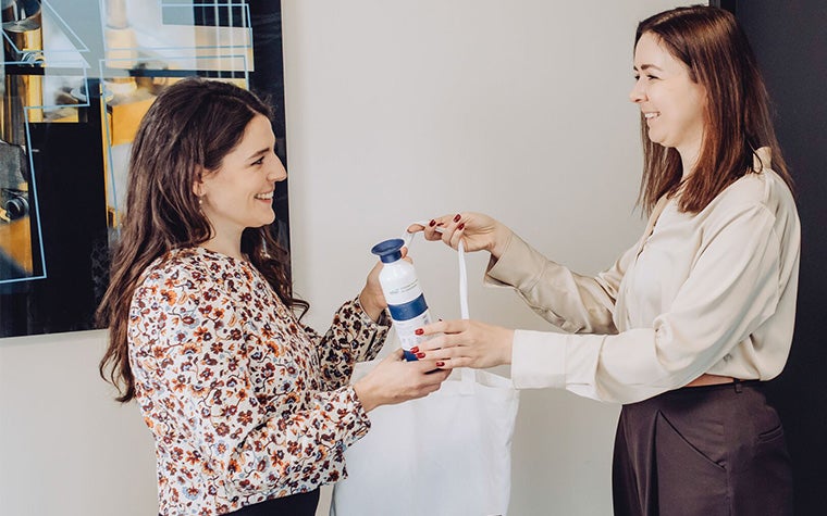 Two women talking in an office environment, exchanging a branded water bottle and a tote bag.