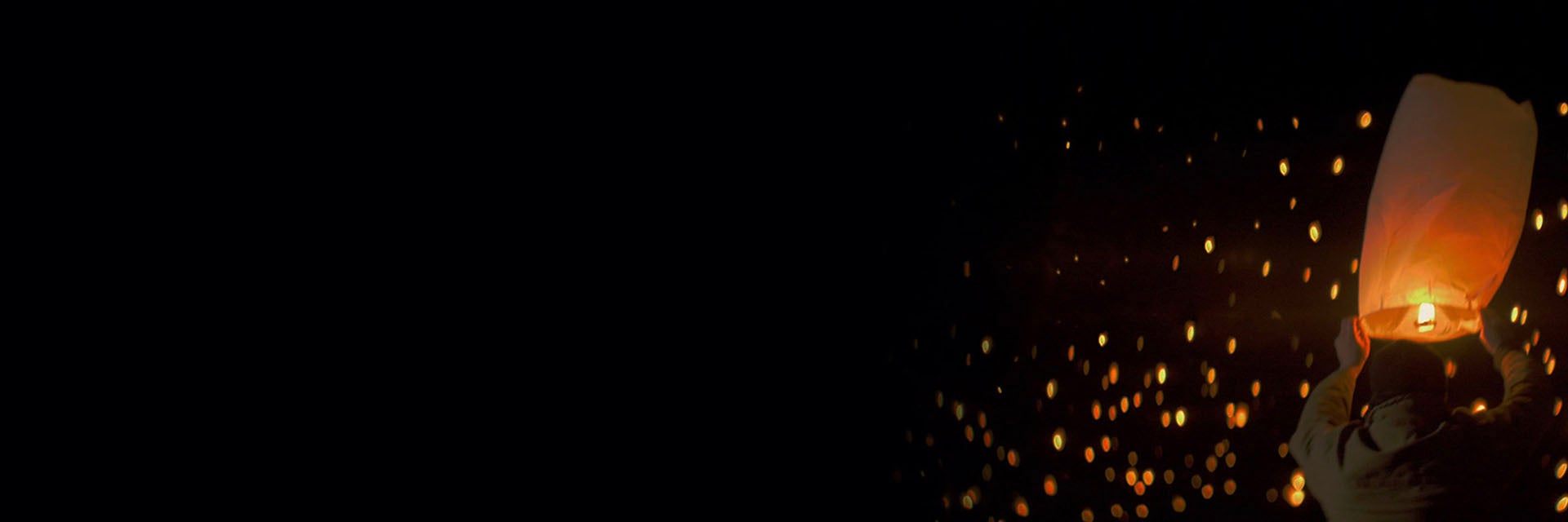 Man holding a lit paper latern overhead with many lights in the distance