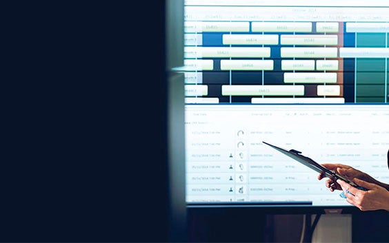 Person holding a clipboard, standing in front of a screen with production management software