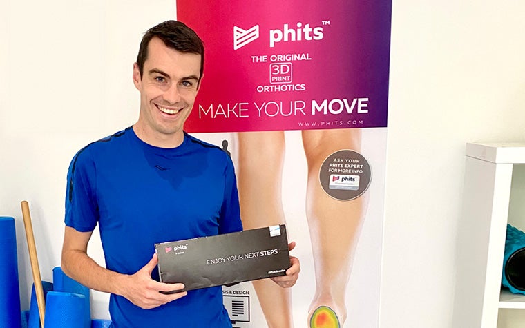 Distance runner Dan Studley stands in front of a phits orthotics banner, holding a box with his custom-made, 3D-printed phits orthotics.