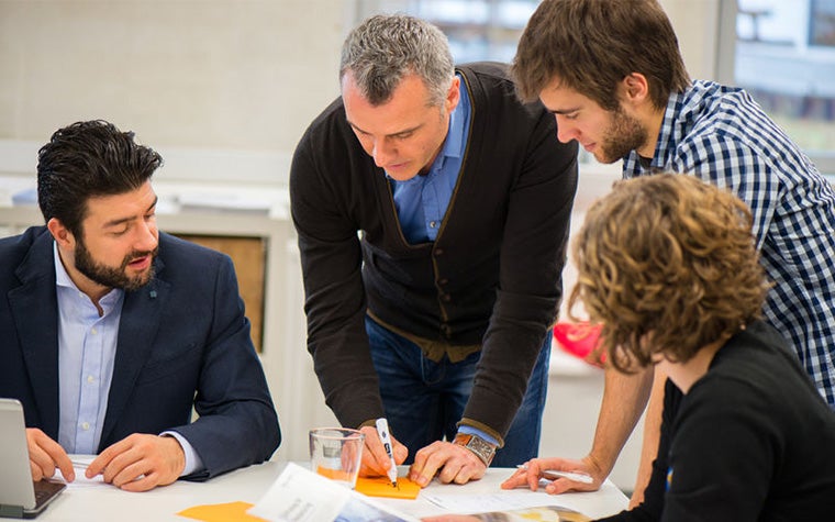Eine Gruppe von Menschen sitzt an einem Tisch und beobachtet, wie ein Mann mit einem Marker auf ein kleines Stück orangefarbenes Papier schreibt.