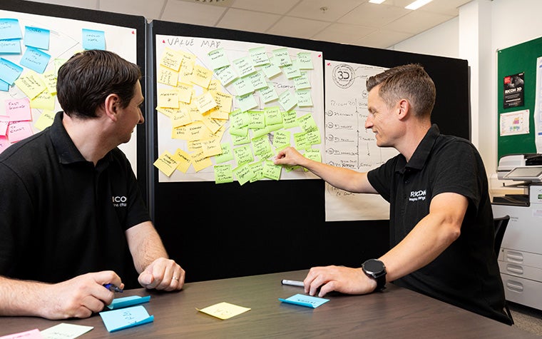 Two Ricoh 3D team members add sticky notes to a value map on a wall