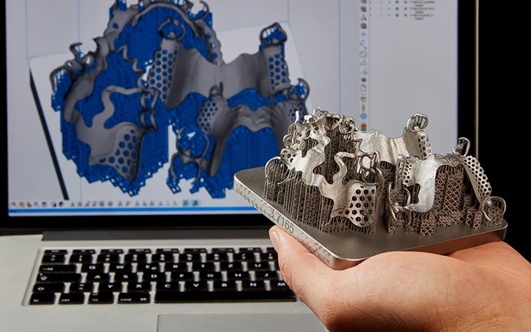 Hand holding a metal 3D-printed part with support structures in front of a screen displaying the design of the part