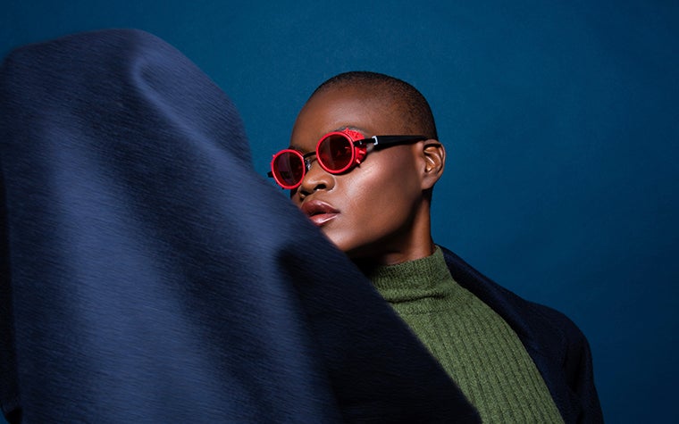 Modèle féminin noir sur fond bleu, regardant hors caméra et portant des lunettes de soleil rouges de la collection NAUTINEW de ReyStudio.