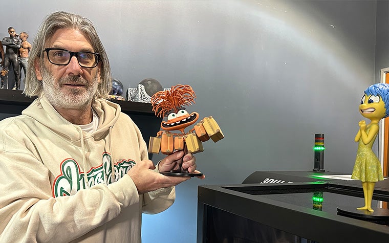 Jason Lopes from Gentle Giant Studios holding a 3D-printed model of Anxiety from Inside Out 2 with another model of Joy from the movie on a nearby 3D printer.