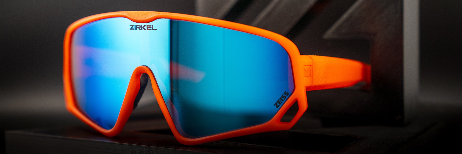 Zirkel's bright orange 3D-printed Teddy Grizz eyewear with a blue visor sitting against a dark grey and black background.