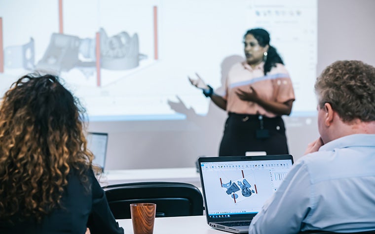 A Materialise Academy trainer standing in front of a projector with Materialise Magics showing a 3D printing build. Two students are sitting at a table in the foreground, looking at laptops showing the same Magics screen