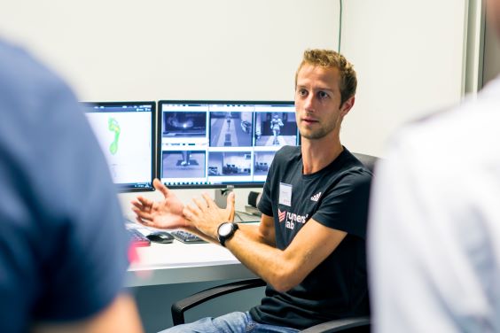 A man in a Runners' Lab t-shirt sits in front of two computer screens, explaining the images of a foot scan to two customers.